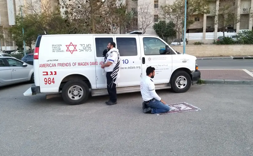 Deux ambulanciers paramédicaux, un juif portant un châle de prière et un musulman utilisant un tapis de prière, prient l'un à côté de l'autre près d'une ambulance Magen David Adom (MDA) à Beersheba, dans le sud d'Israël, le 24 mars 2020 (crédit photo: MOHAMD ALNBARE / MDA SPOKESPERSON)